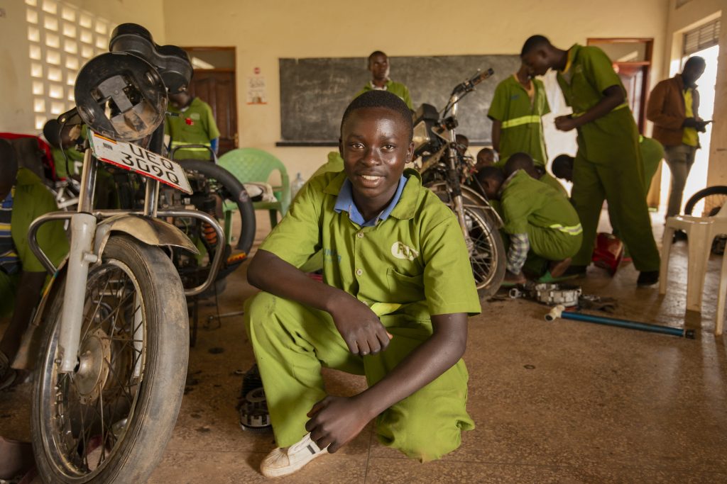 A boy infront of a class while others are looking at a motorcycle in the back