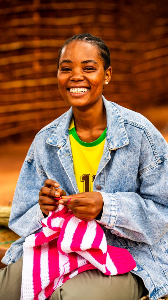 A smiling woman sitting on a mat crocheting a piece of cloth